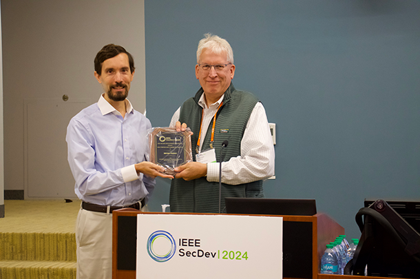 Bryan Parno (left) receives award plaque from Greg Shannon behind a podium with IEEE SecDev 2024 sign