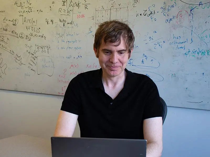 A man sits at a laptop, in front of a dry erase board covered in complicated equations.